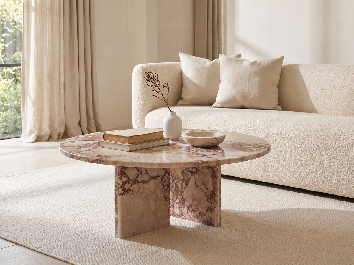 Paestum Rosa Marble Coffee Table styled with books, a vase and a stone bowl in front of a cream sofa in a bright plaster room