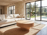 Pesaro Ash Veneer Fluted Pedestal Coffee Table seen from the side, on a jute rug in front of a linen sofa in a poolside villa