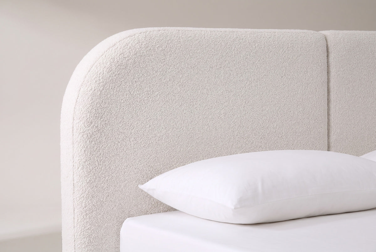 The rounded top corner of the Pietrabbondante headboard in ivory boucle, with the seam between the two panels and a white pillow below