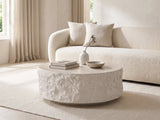 Piombino Faux Travertine Fiberglass Drum Coffee Table styled with books, a vase and a stone bowl in front of a cream sofa in a bright plaster room