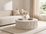 Piombino Faux Travertine Fiberglass Drum Coffee Table seen from the side, styled with books, a vase and a stone bowl in front of a cream sofa in a bright plaster room