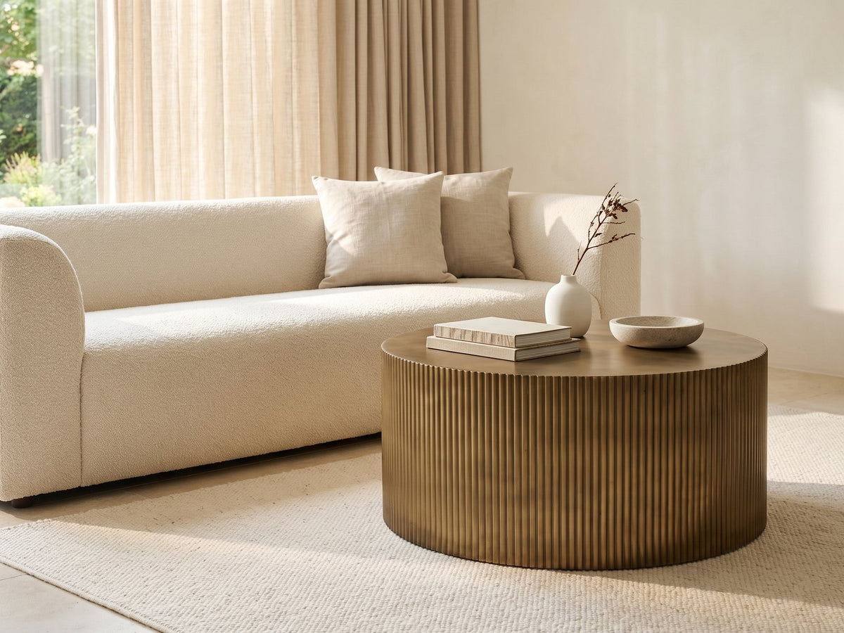 Pitigliano Brass-Finish Fluted Drum Coffee Table styled with books, a vase and a stone bowl in front of a cream sofa in a bright plaster room