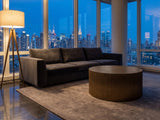 Pitigliano Brass-Finish Fluted Drum Coffee Table seen from the side, on a wool rug in front of a velvet sofa in a city penthouse at blue hour