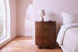 Ploaghe chestnut mango oval nightstand with a ceramic lamp beside a bed in a Los Angeles bungalow bedroom with a blush pink plaster wall and terracotta floor tiles