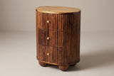 The Ploaghe oval three-drawer nightstand in chestnut mango wood with fine vertical reeds and brass knobs, three-quarter view on a light studio backdrop