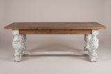 The Policoro dining table, front view of the long side on a light studio backdrop: a fir apron above four carved urn legs and one stretcher