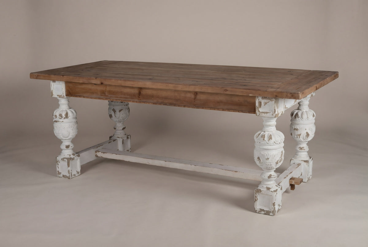The Policoro dining table with a weathered fir plank top and four carved antique-white urn legs, three-quarter view on a light studio backdrop