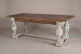 The Policoro dining table with a weathered fir plank top and four carved antique-white urn legs, three-quarter view on a light studio backdrop