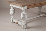 A near corner of the Policoro dining table: the fir top, a square block under it, a carved antique-white urn leg on a square block foot, the end rail with its small wooden key and the stretcher