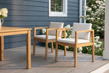 Two Pollina acacia dining armchairs on a grey porch deck beside an acacia dining table, a slate-blue shiplap wall and white hydrangeas behind (table not included)