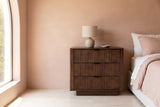 Pula reeded walnut three-drawer nightstand with a ceramic lamp beside a bed in a Los Angeles bedroom with a blush-pink lime plaster wall and an arched window