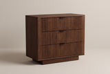 The Pula three-drawer nightstand in dark walnut with fully reeded drawer fronts and black tab pulls, three-quarter view on a light studio backdrop