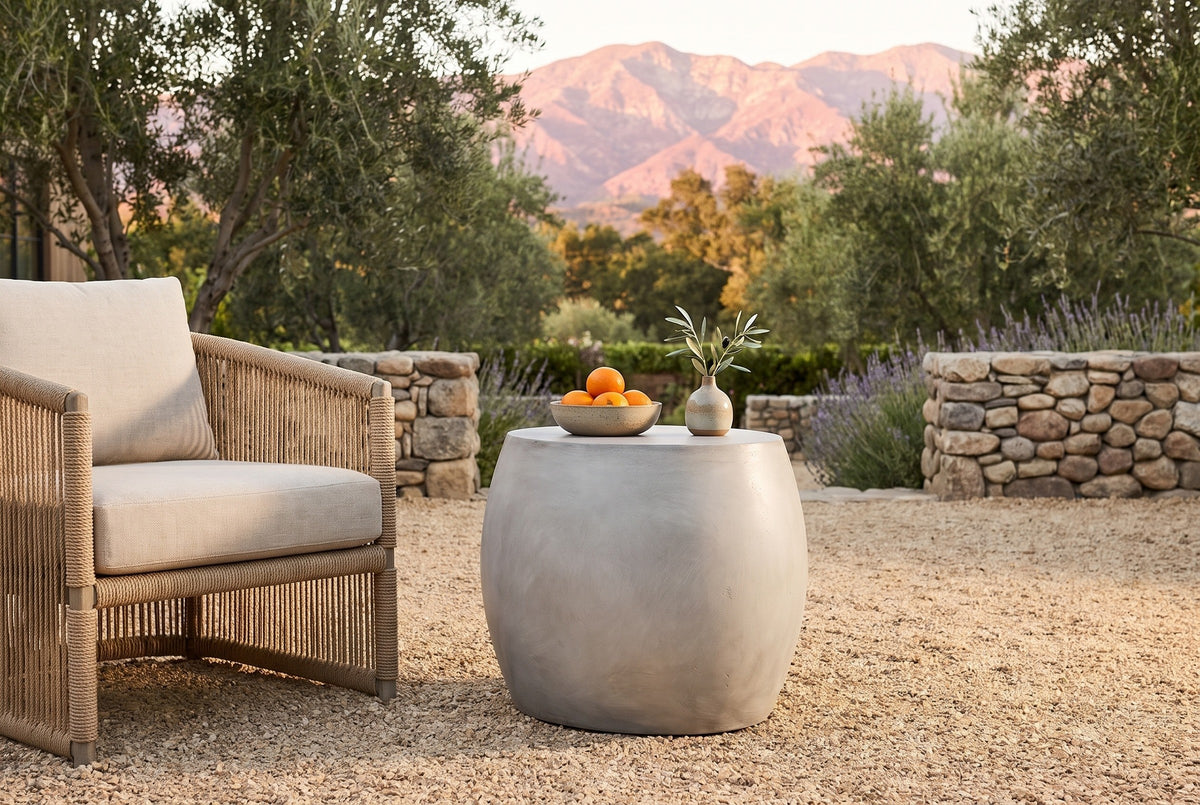 Racalmuto concrete barrel side table with a bowl of oranges and a small vase of olive on a gravel terrace in Ojai, California at golden hour, a river-stone wall, olive trees and the mountains behind (chair and decor not included)