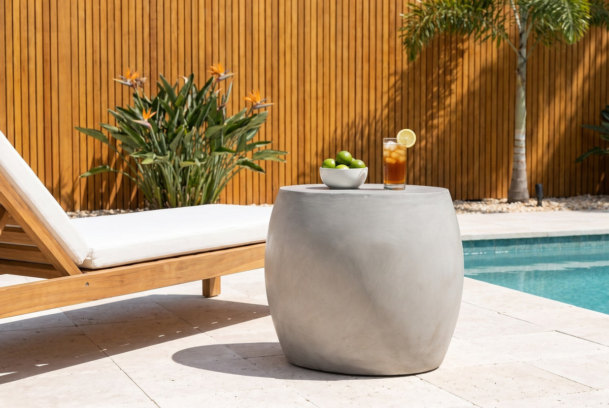 Racalmuto light grey concrete barrel side table with a bowl of limes and a glass of iced tea beside a teak lounger on a limestone pool terrace in Sarasota, a cedar slat wall behind (lounger and decor not included)
