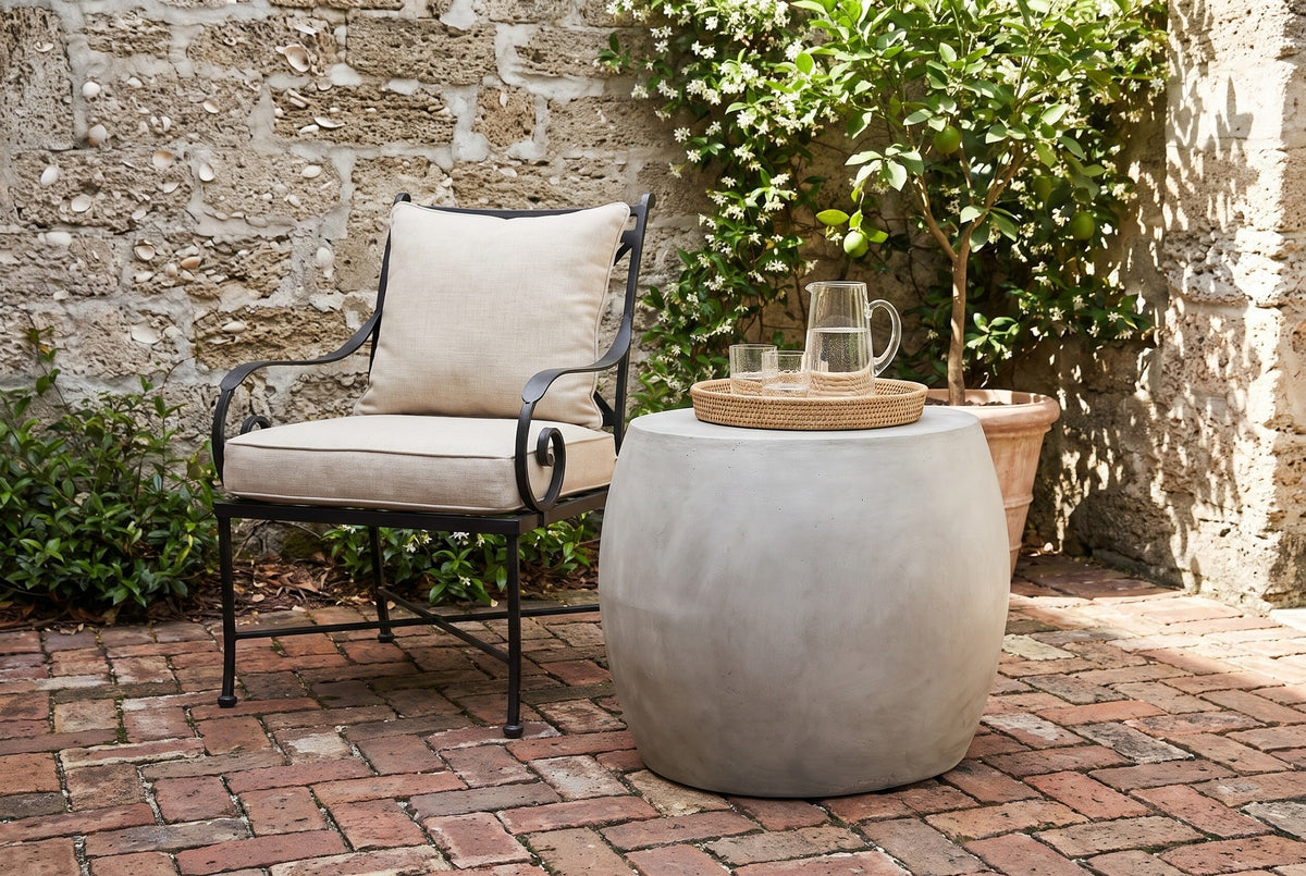 Racalmuto concrete barrel side table with a glass pitcher and glasses on a woven tray in a St. Augustine courtyard, a coquina shell-stone wall, star jasmine and an old brick floor (chair and decor not included)