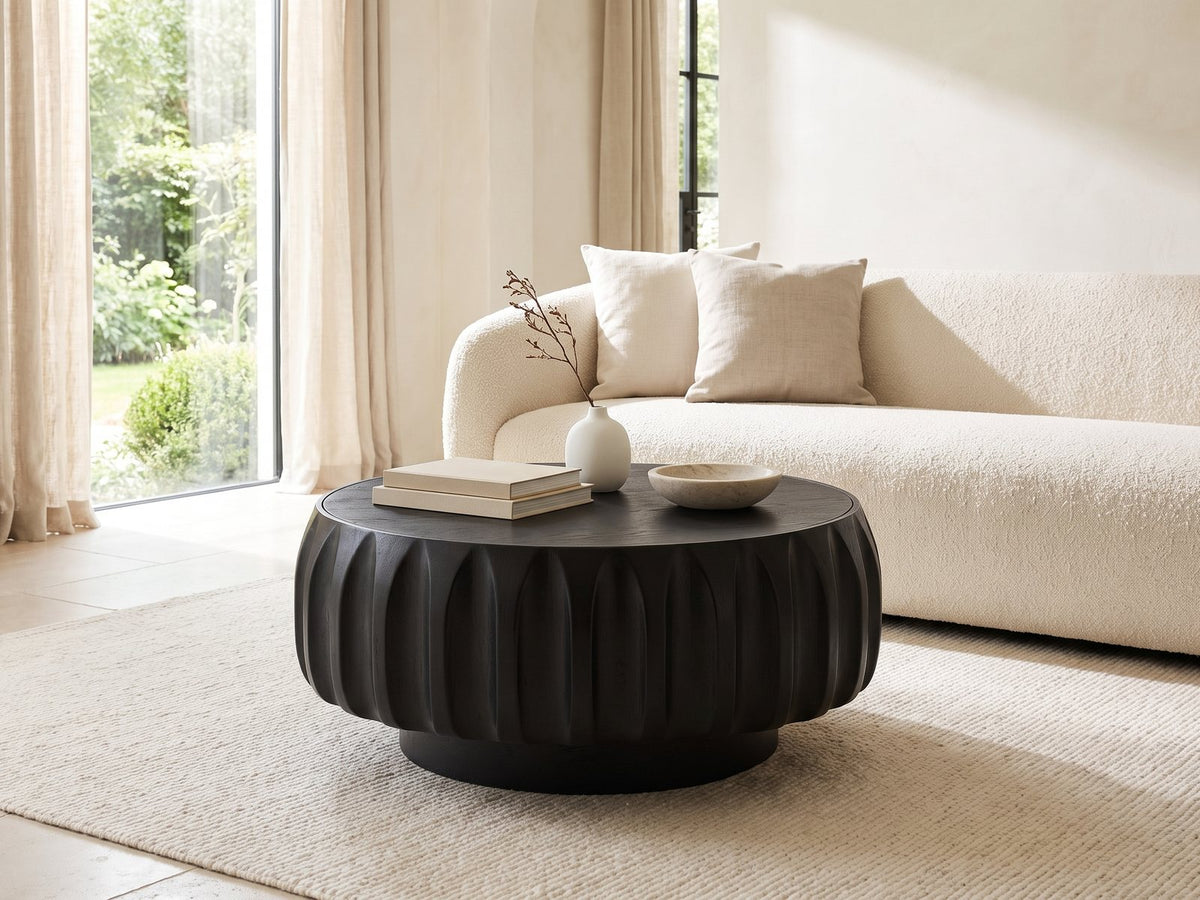 Radda Black Carved Wood Drum Coffee Table styled with books, a vase and a stone bowl in front of a cream sofa in a bright plaster room