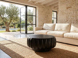 Radda Black Carved Wood Drum Coffee Table seen from the side, on a jute rug in front of a linen sofa in a poolside villa