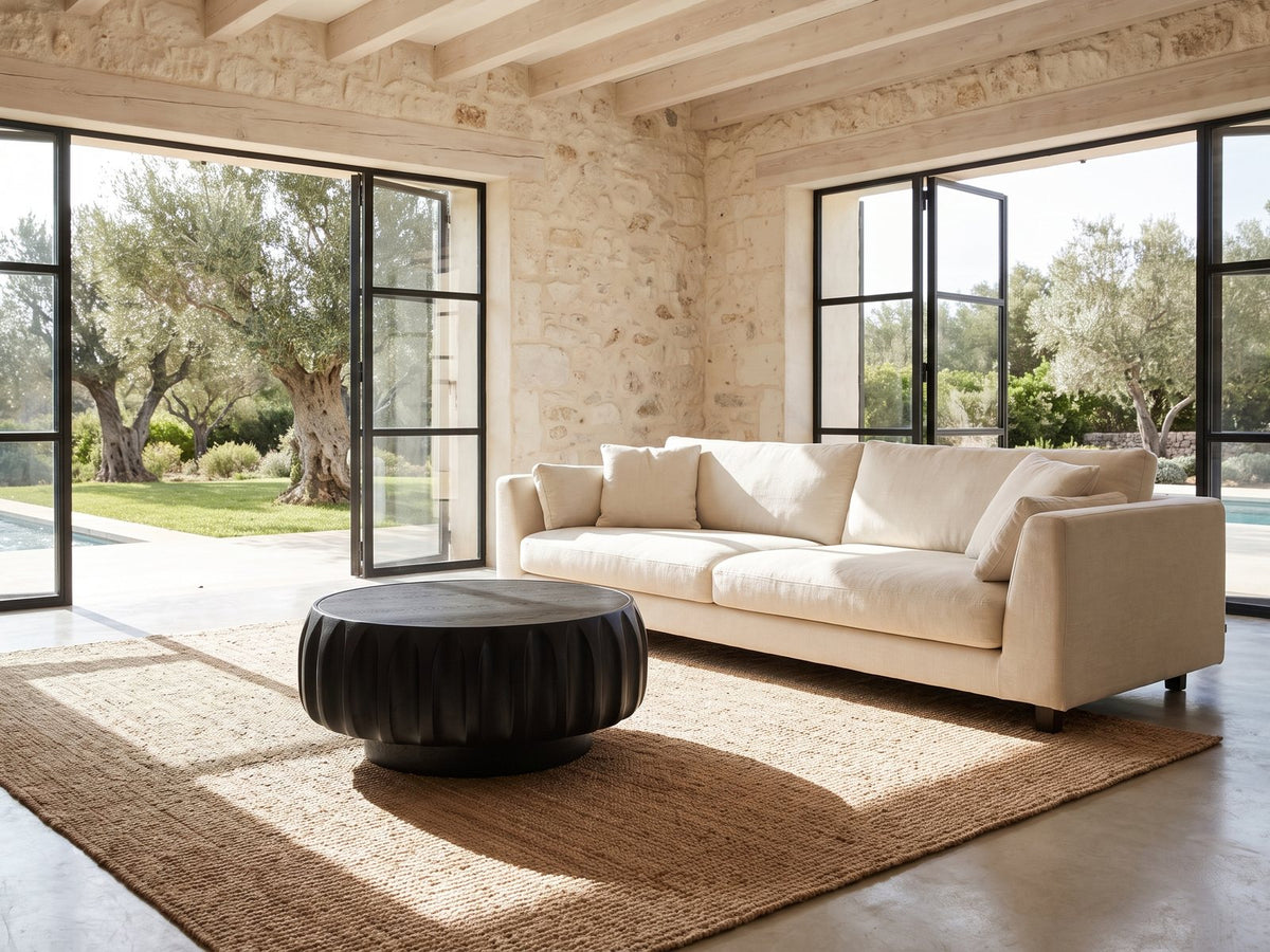 Radda Black Carved Wood Drum Coffee Table on a jute rug in front of a linen sofa in a poolside villa
