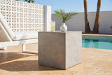 Realmonte light grey concrete cube side table with a glass of lemonade and a white vase beside a white lounge chair on a travertine pool patio in Palm Springs, a white breeze-block wall behind (chair and decor not included)