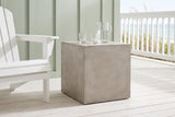 Realmonte concrete cube side table with a water jug and glasses on a beach-house porch in Seaside, Florida, a sage-green board wall and the Gulf beyond (chair and decor not included)