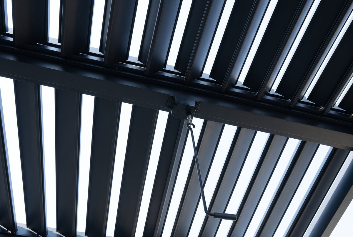 Looking up at the Regalbuto roof: black aluminum louvers tilted open under the sky, a cross beam, the gearbox and the crank hook