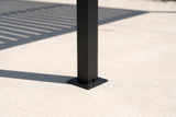 The foot of a Regalbuto black aluminum post on its square base plate on a pale concrete patio, louver shadows beside it
