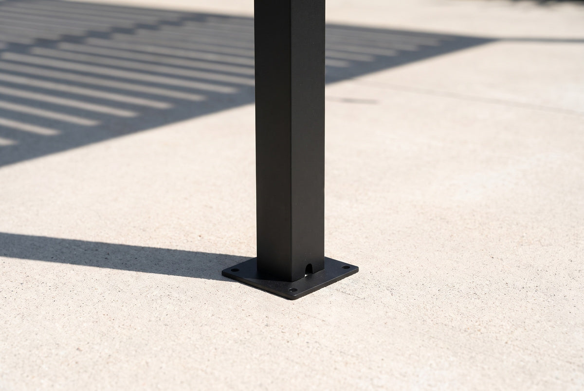 The foot of a Regalbuto black aluminum post on its square base plate on a pale concrete patio, louver shadows beside it