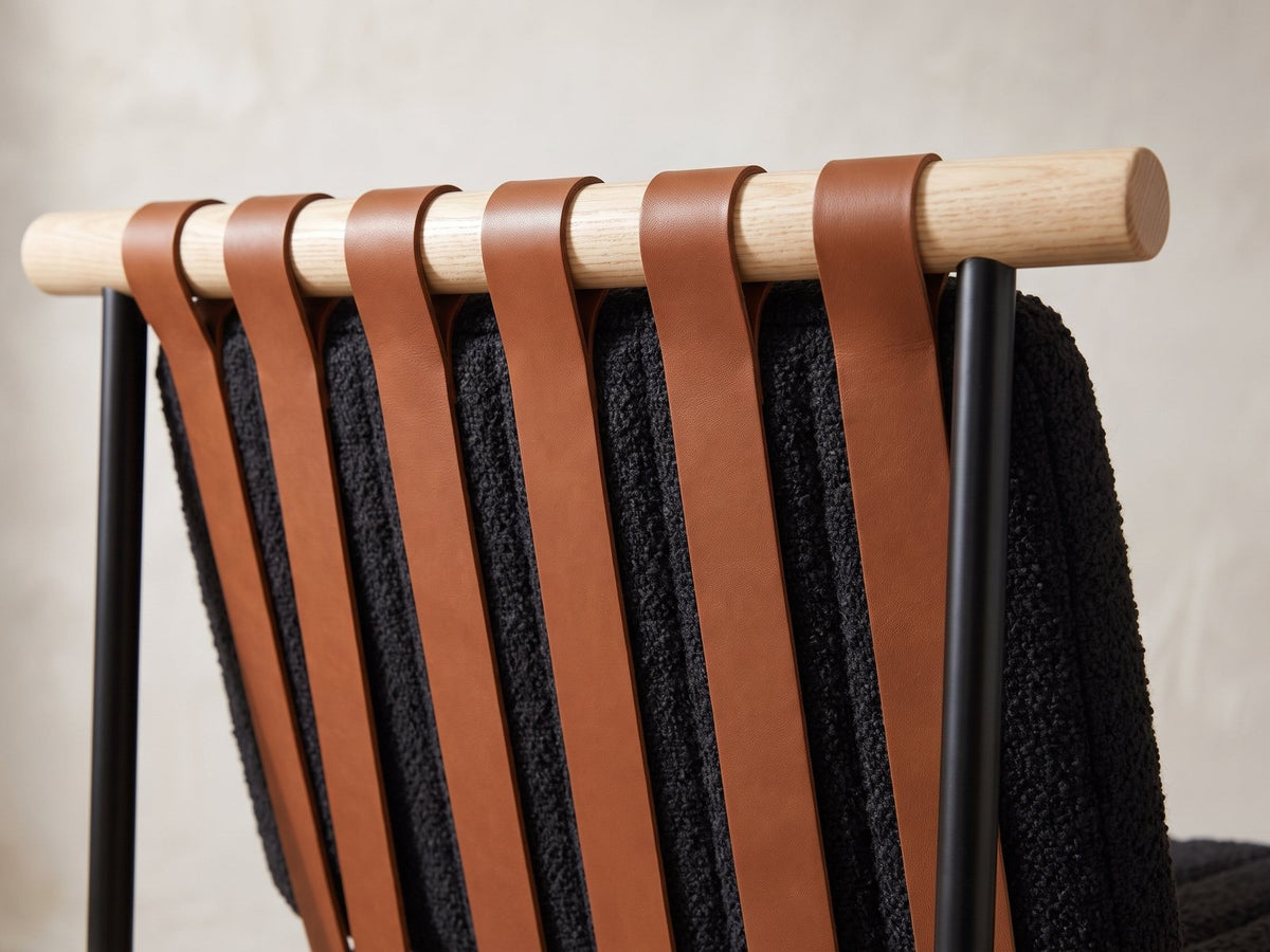 Close-up of the Rende 59-Inch Round Black Oak Hourglass Dining Set with 6 Chairs: cognac straps over a pale oak rail on the back of a black boucle chair
