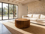Rivoli 30-Inch Round Carved Wood Drum Coffee Table on a jute rug in front of a linen sofa in a poolside villa