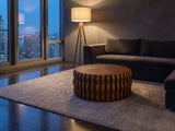 Rivoli 30-Inch Round Carved Wood Drum Coffee Table on a wool rug in front of a velvet sofa in a city penthouse at blue hour