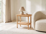 Rutigliano Braided Rattan Two-Tier Side Table with a lamp and a ceramic bowl beside a cream boucle armchair in a bright plaster room