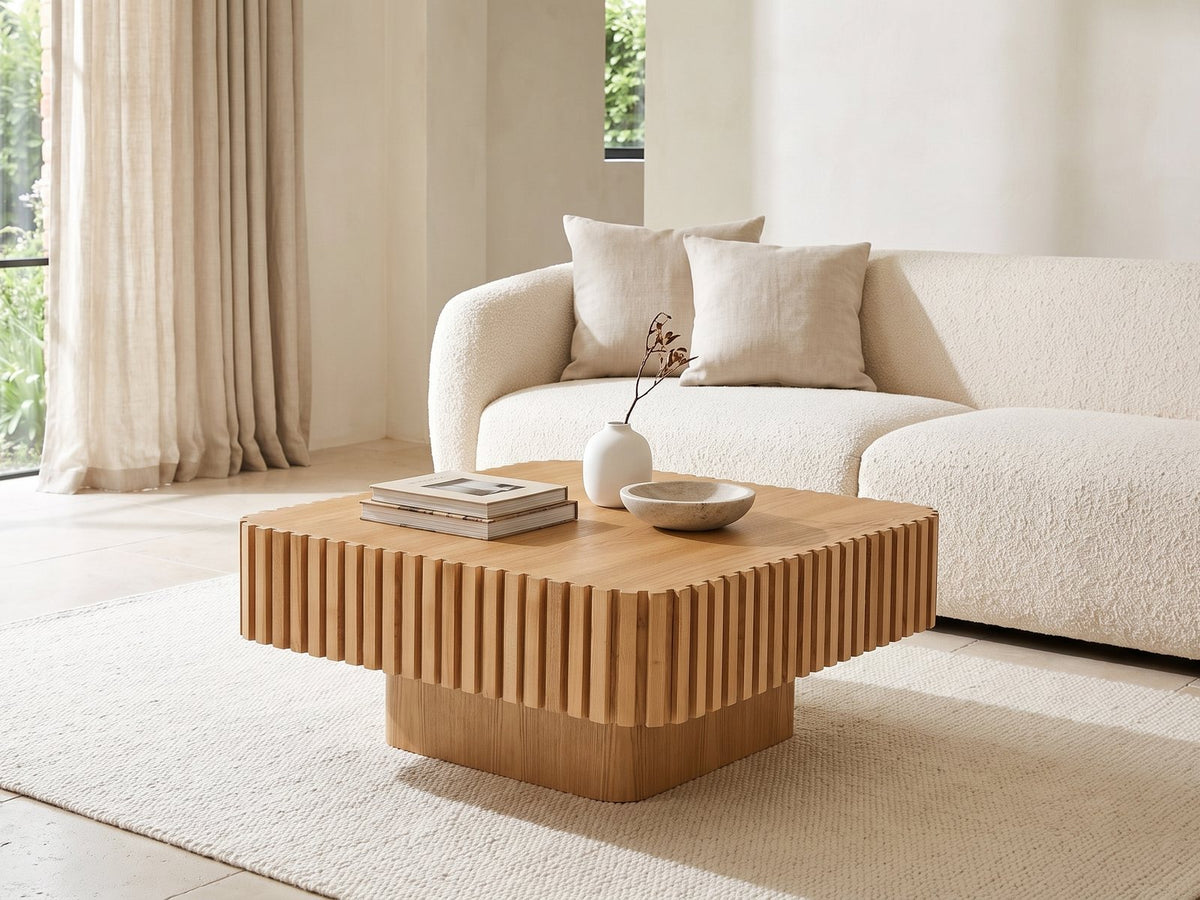 Saluzzo 35-Inch Square Slatted Natural Wood Veneer Coffee Table seen from the side, styled with books, a vase and a stone bowl in front of a cream sofa in a bright plaster room