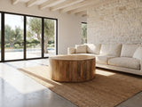 Sansepolcro Natural Mango Drum Coffee Table seen from the side, on a jute rug in front of a linen sofa in a poolside villa
