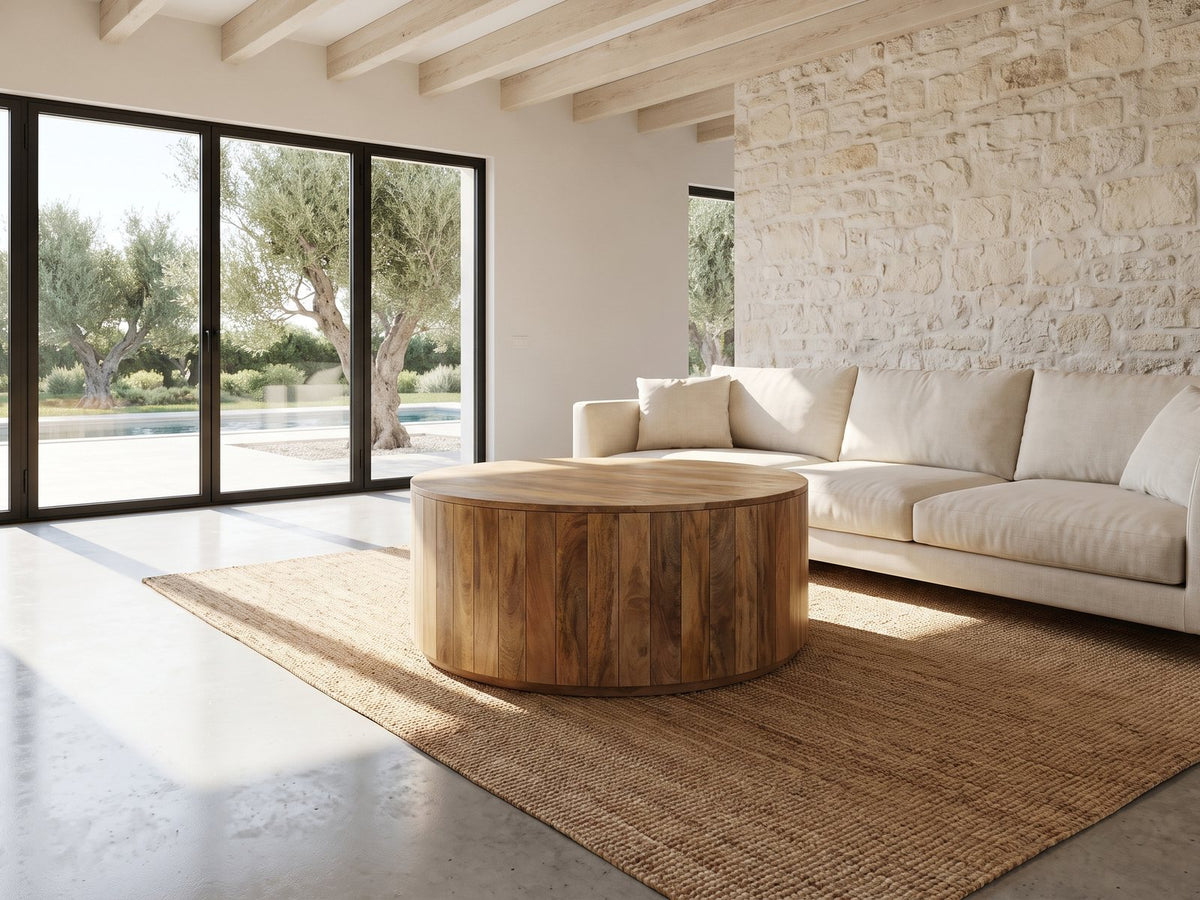 Sansepolcro Natural Mango Drum Coffee Table seen from the side, on a jute rug in front of a linen sofa in a poolside villa