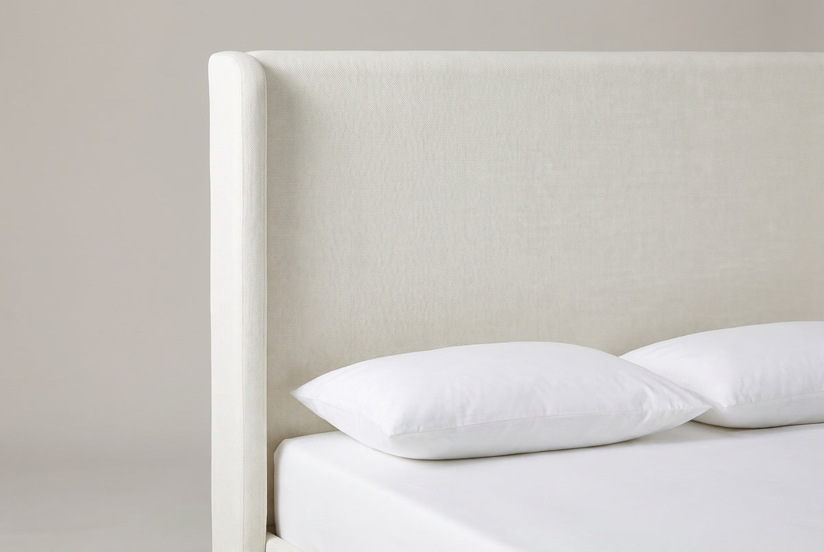 One side of the Scanno headboard: a tall flat ivory canvas panel with a slim rounded wing running down beside it, above a white mattress with two white pillows