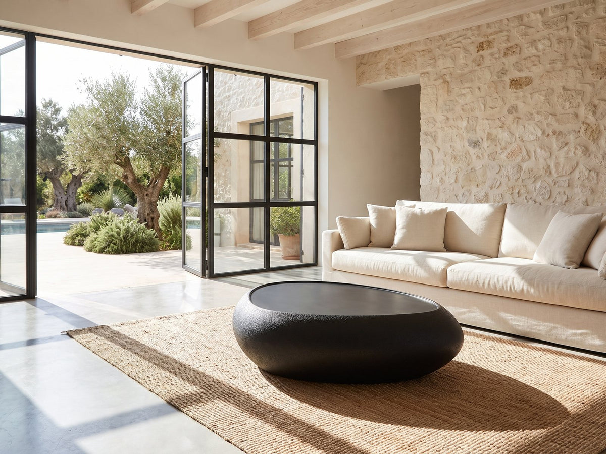Scicli Black Fiberglass Pebble Coffee Table seen from the side, on a jute rug in front of a linen sofa in a poolside villa