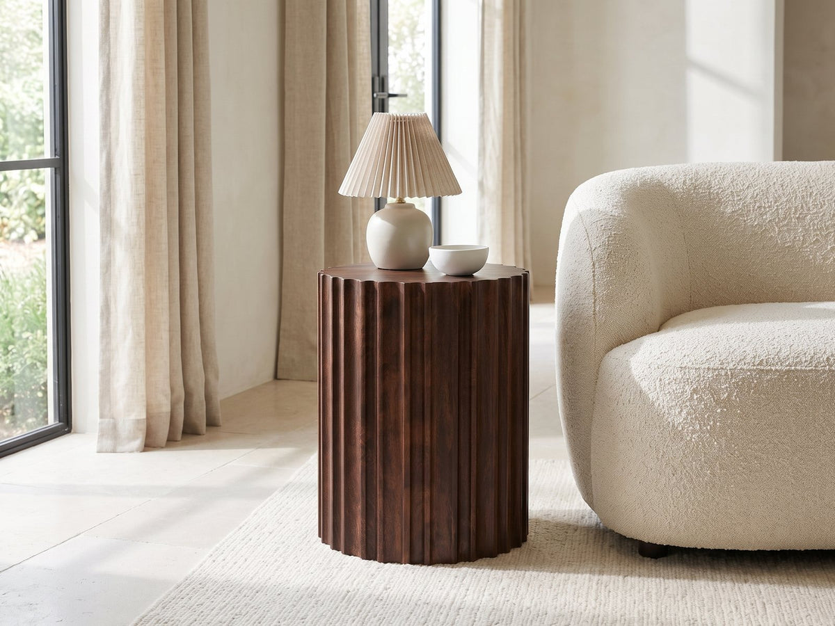 Senise Walnut Mango Fluted Drum Side Table with a lamp and a ceramic bowl beside a cream boucle armchair in a bright plaster room