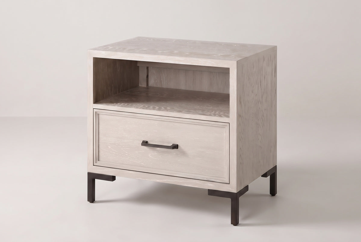 The Sennori nightstand in pale cerused oak with an open shelf, one drawer and dark bronze legs, three-quarter view on a light studio backdrop