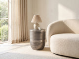 Sepino Antique Silver Metal Drum Side Table with a lamp and a ceramic bowl beside a cream boucle armchair in a bright plaster room