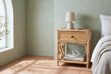 Sinnai rattan nightstand with a ceramic lamp and a folded throw on its shelf beside a bed in an Ojai bedroom with a sage plaster wall and terracotta tiles