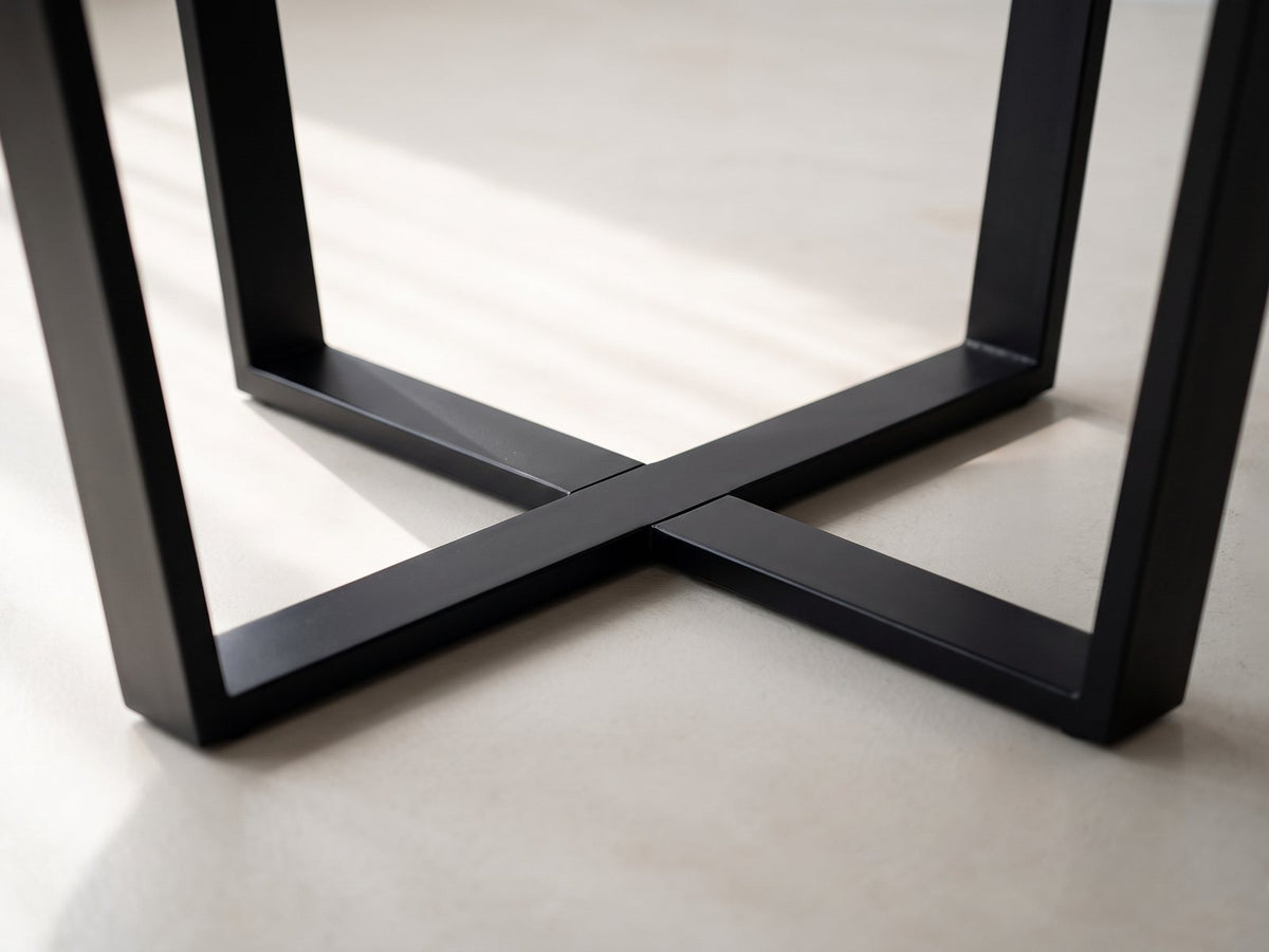 Close-up of the Soveria 63-Inch Round Black Acacia Sunburst Dining Table: the black iron base: two U-shaped frames crossing in a flat X on the floor