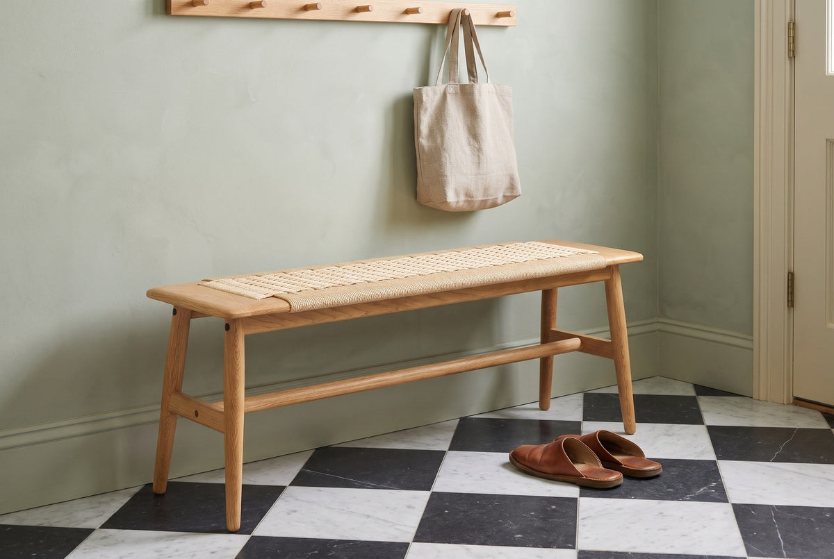 Sperlonga oak bench with a paper cord seat along a sage-green plaster wall in a Charleston townhouse entry, with a wooden peg rail, a linen tote bag and a black and white marble floor (bag and shoes not included)