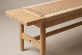 One end of the Sperlonga bench: the flat oak end block with a rounded corner, the paper cord seat beside it, the oak side rail below, a round tapered leg and the flat stretcher with the long round rail