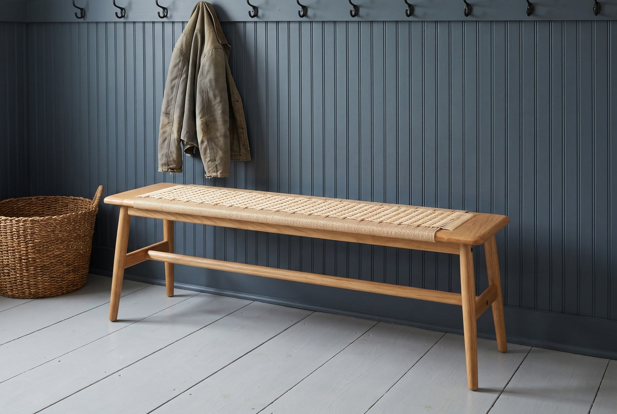 Sperlonga oak bench with a paper cord seat along a slate blue-grey beadboard wall in a Vermont mudroom with black iron hooks, a canvas jacket, a woven basket and a pale grey painted floor (jacket and basket not included)
