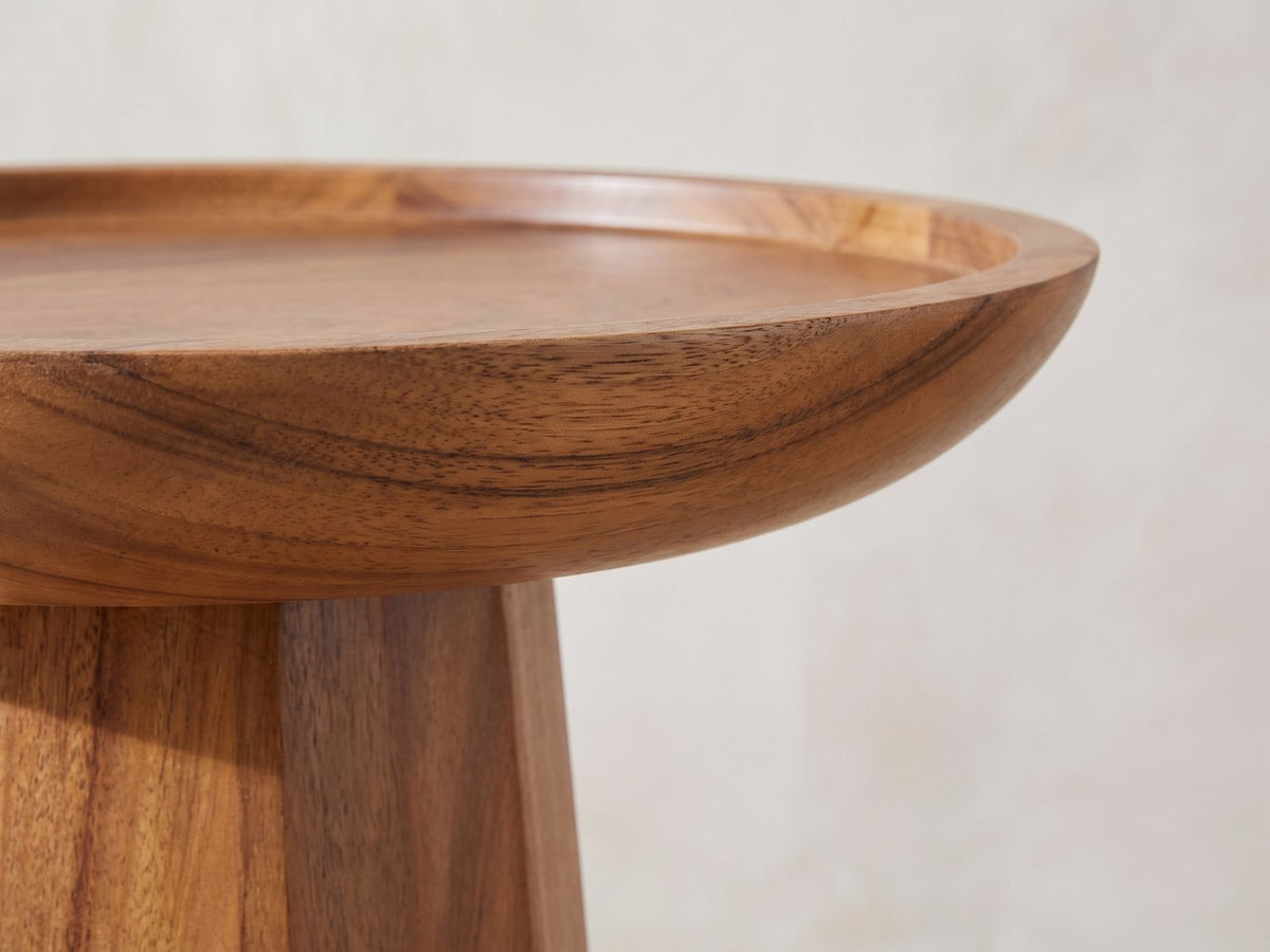 Close-up of the Sternatia Cognac Acacia Pedestal Side Table: the raised tray rim and curved underside over the faceted pedestal