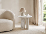 Stintino White Marble Round Side Table with a lamp and a ceramic bowl beside a cream boucle armchair in a bright plaster room