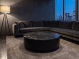 Taormina Black Marble Drum Coffee Table seen from the side, on a wool rug in front of a velvet sofa in a city penthouse at blue hour