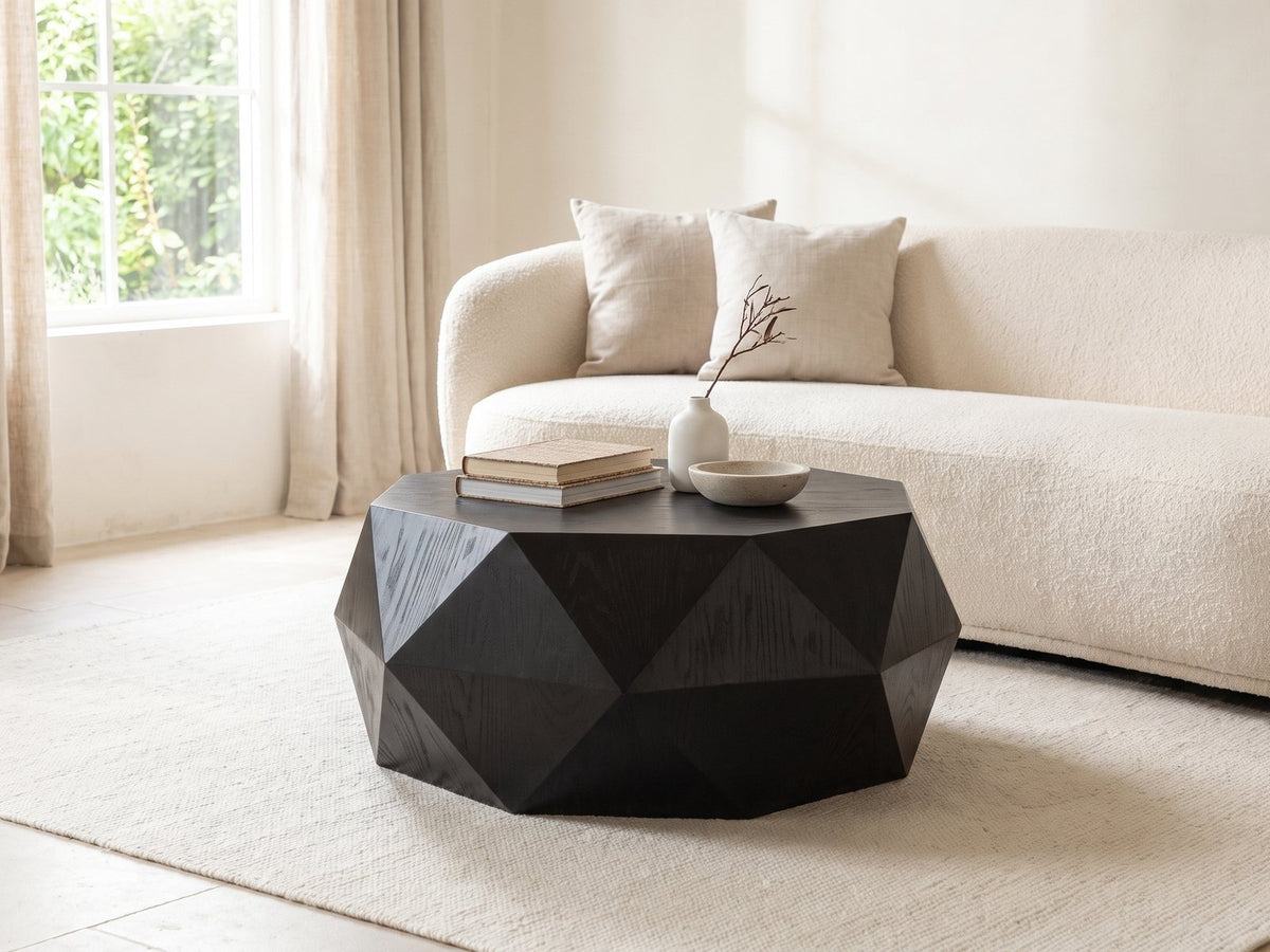 Taranto Black Faceted Drum Coffee Table styled with books, a vase and a stone bowl in front of a cream sofa in a bright plaster room