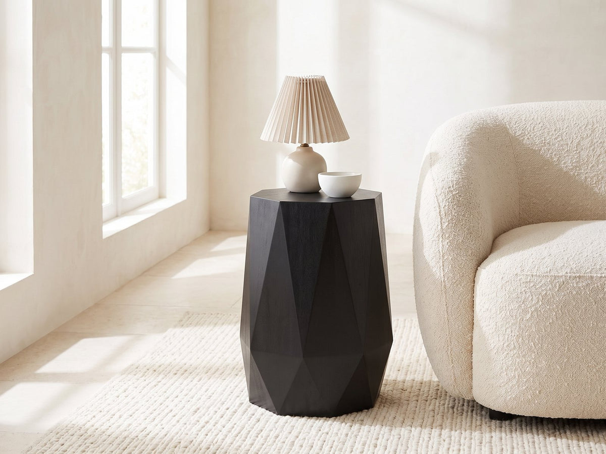 Teglio Black Acacia Faceted Octagon Side Table with a lamp and a ceramic bowl beside a cream boucle armchair in a bright plaster room