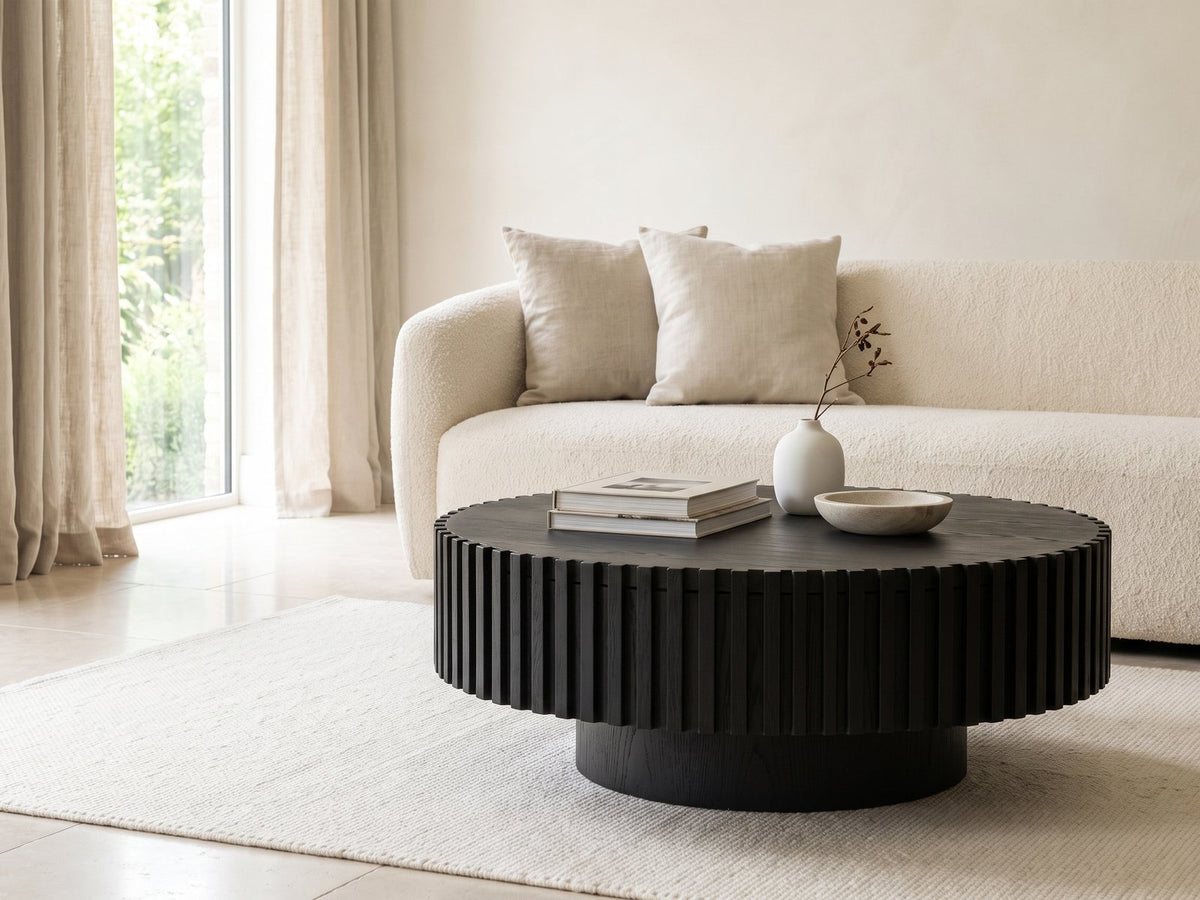 Terni Black Wood Veneer Fluted Pedestal Coffee Table styled with books, a vase and a stone bowl in front of a cream sofa in a bright plaster room
